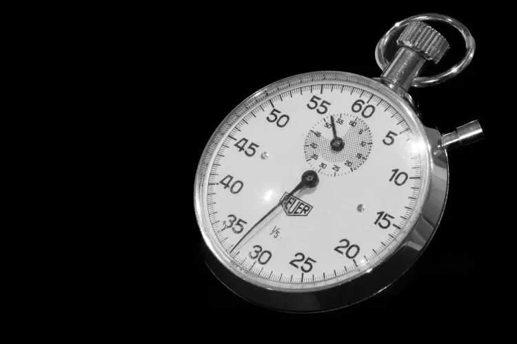 Eine silberne mechanische Stoppuhr mit weißem Zifferblatt und Minutenskala schräg vor schwarzem Hintergrund, Schwarz-Weiß-Aufnahme
