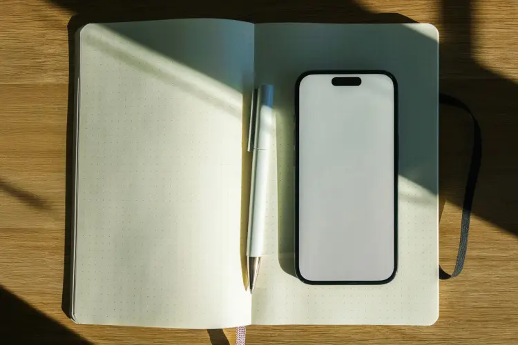 An open dot-grid notebook lying in sunlight on a wooden desk, with a ballpoint pen and a smartphone with a blank screen resting on the page