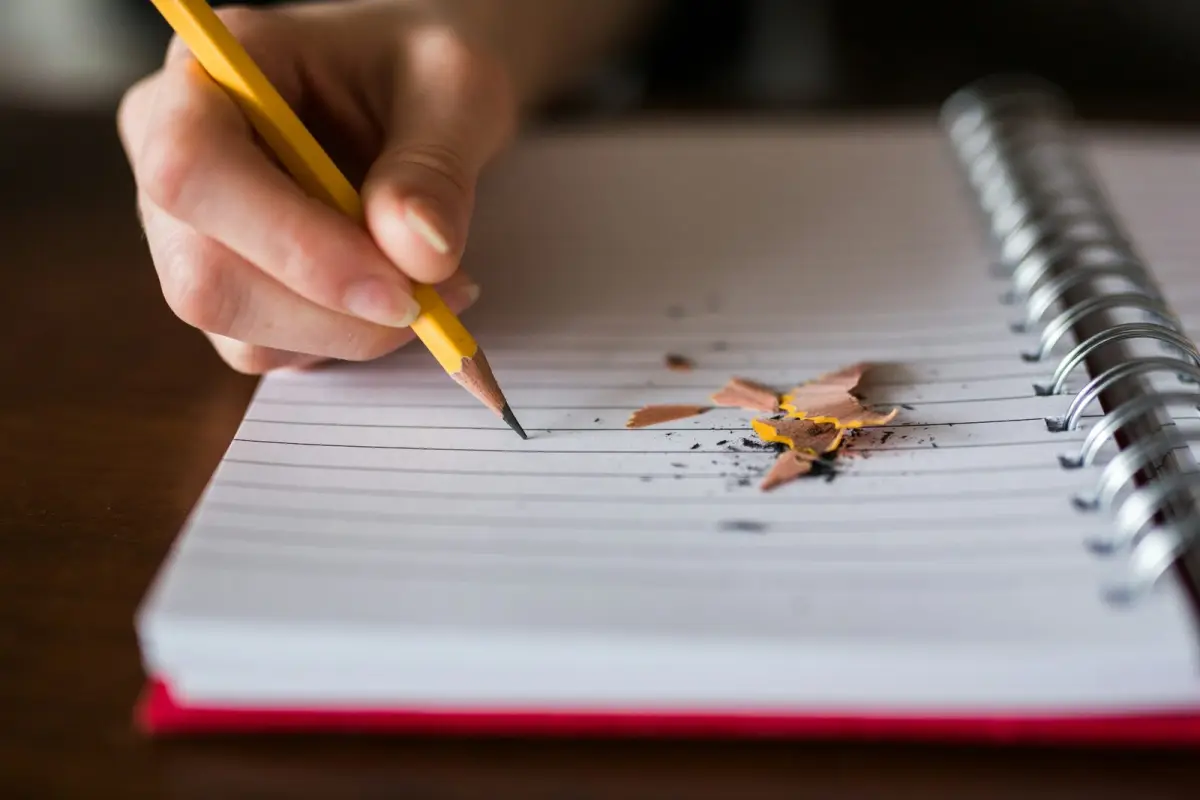 A hand setting a sharpened pencil to the blank ruled page of a notebook, pencil shavings beside it