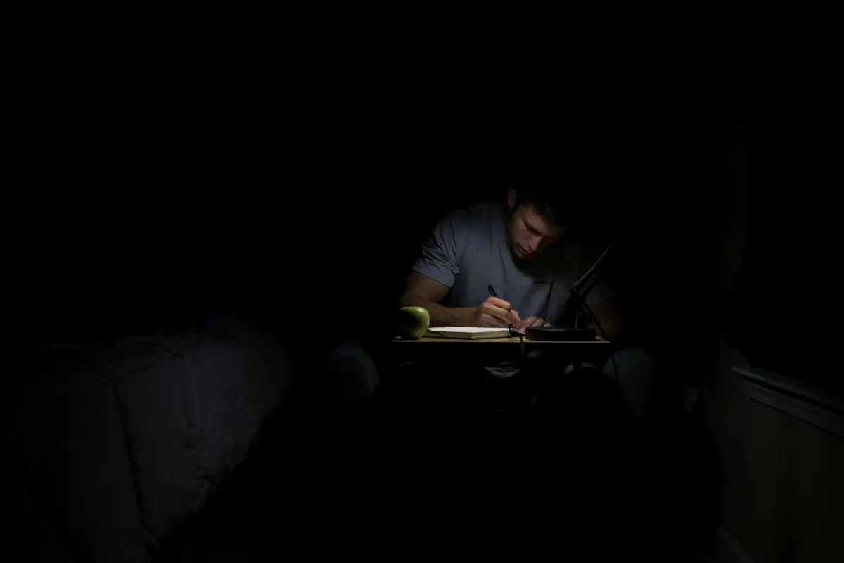 A man writing at a small desk in a darkened room, the desk lamp the only source of light