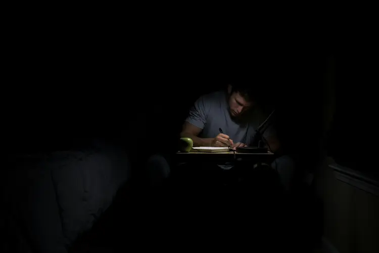 A man writing at a small desk in a darkened room, the desk lamp the only source of light