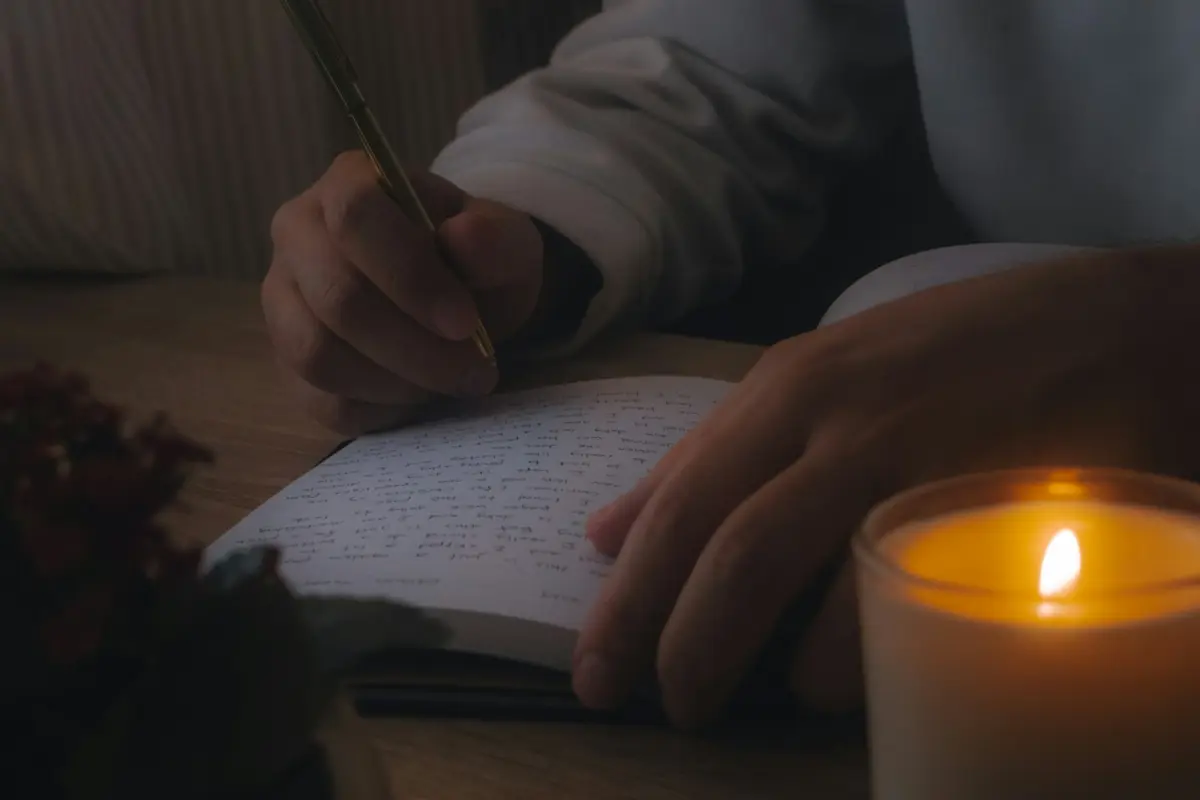 Someone writing with a pen in an open notebook, a candle burning in a glass beside it