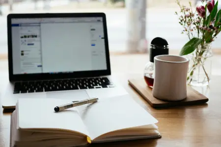 Ein aufgeklappter Laptop auf einem Holztisch am Fenster, davor ein offenes Notizbuch mit Stift, daneben ein weißer Becher und eine Vase mit Blumen