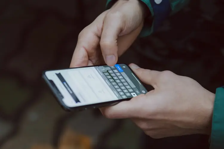 Two hands hold an iPhone sideways while a finger taps the on-screen keyboard, with a dark blurred background behind them