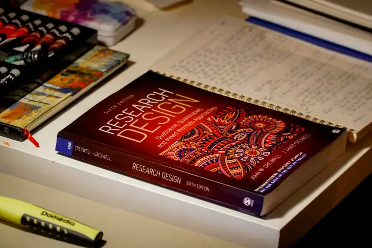 A research methods textbook with a red cover lies on a pale desk next to a spiral notebook full of handwritten notes, with a yellow highlighter in the foreground