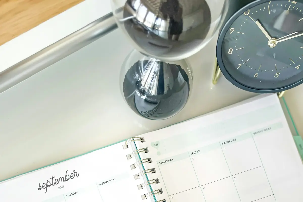 An hourglass with black sand and a dark blue desk clock stand on a pale tray next to an open monthly planner showing the September page