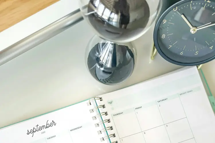 An hourglass with black sand and a dark blue desk clock stand on a pale tray next to an open monthly planner showing the September page