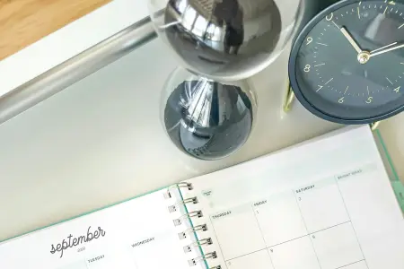 Eine Sanduhr mit schwarzem Sand und eine dunkelblaue Tischuhr stehen auf einem hellen Tablett, davor liegt ein aufgeschlagener Monatsplaner mit der Seite für September