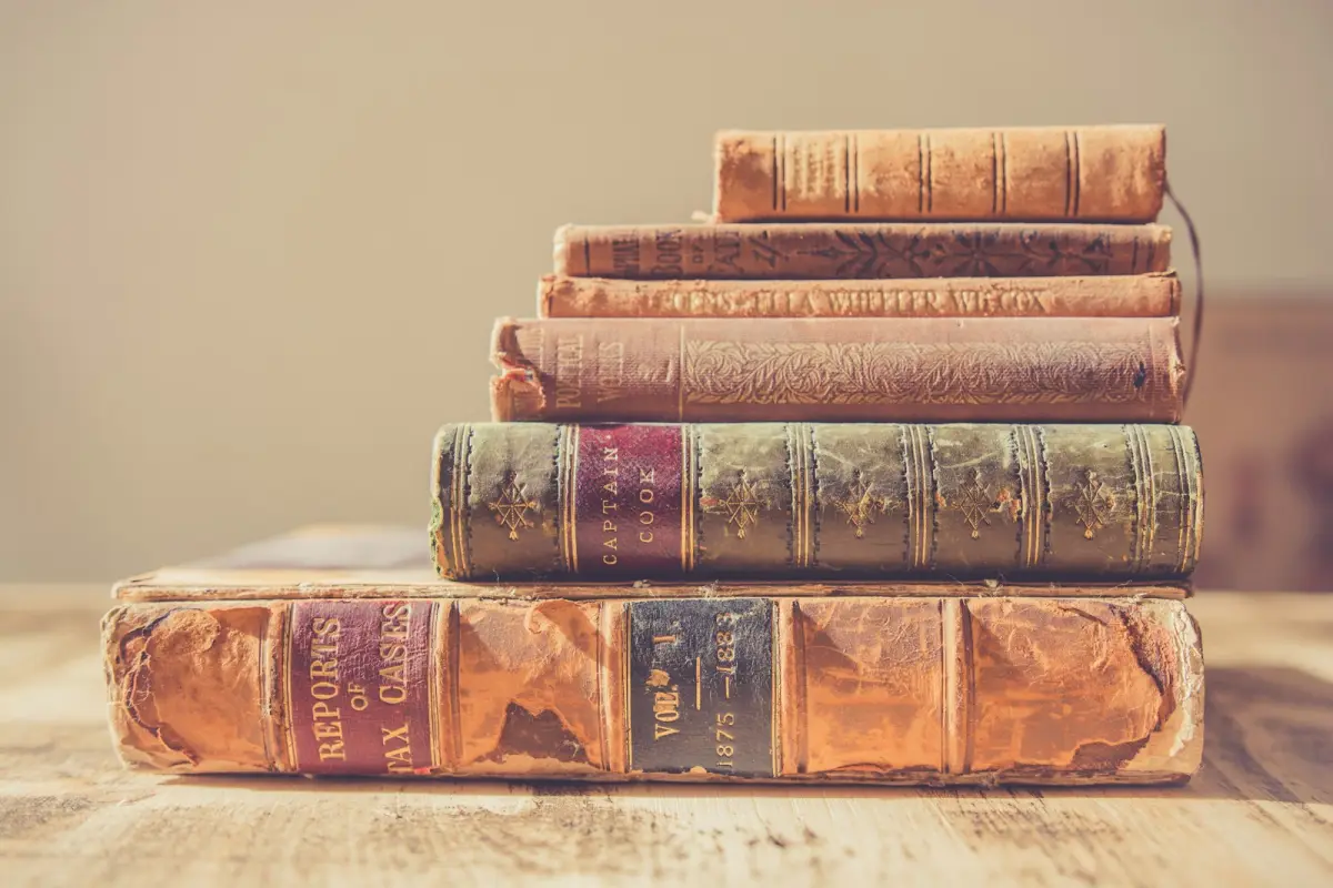 Six old leather-bound books of different sizes stacked on a wooden table, the largest volume at the bottom and the smallest on top